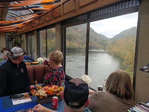 A group of people inside a train car, enjoying a scenic view of a river and autumn foliage through large windows.