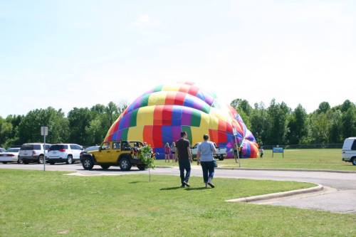 A couple walks towards a colorful hot air balloon being inflated in a grassy field with parked cars nearby.