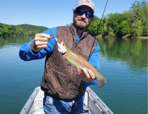 A man in a blue shirt and brown vest holds a rainbow trout while fishing on a calm lake surrounded by greenery.