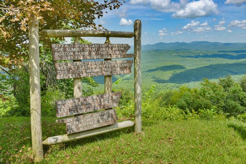 A wooden sign with information about Buffalo River Canyon and the Ozark Mountains, surrounded by lush greenery and blue skies.