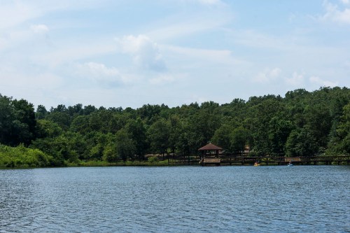 A serene lake surrounded by lush greenery, with a wooden gazebo and a clear blue sky above.