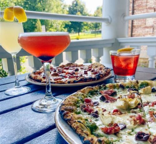 Three cocktails and two pizzas on a table outdoors, with a sunny park view in the background.
