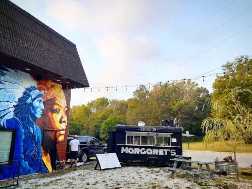 A food truck named "Margaret's" beside a mural of a Native American and a woman, with trees and a cloudy sky in the background.