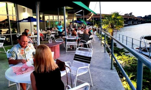 Outdoor dining area by the water, with people enjoying meals at tables under umbrellas.