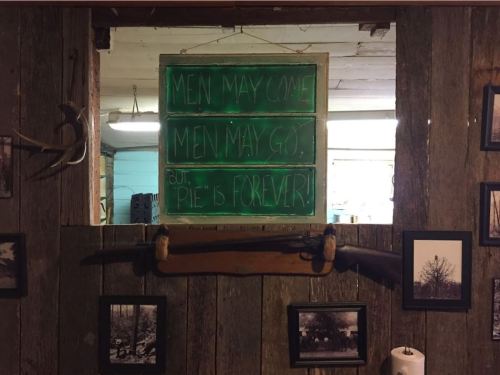 A rustic wooden wall with a neon sign reading "Men may come, men may go, but pie is forever!" and framed photos.