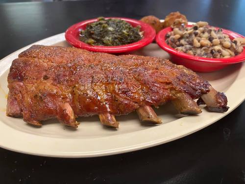A plate of glazed ribs served with collard greens, black-eyed peas, and cornbread muffins in red bowls.