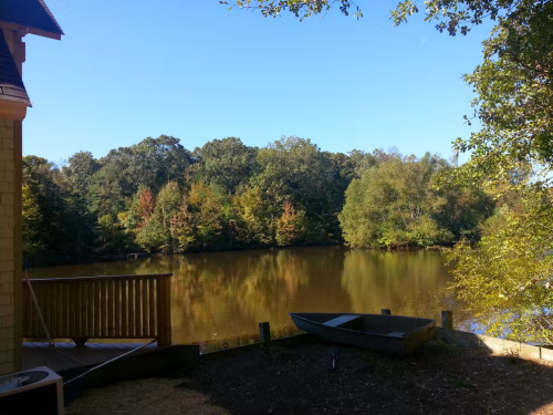A serene view of a calm lake surrounded by trees, with a small boat on the shore and a clear blue sky above.