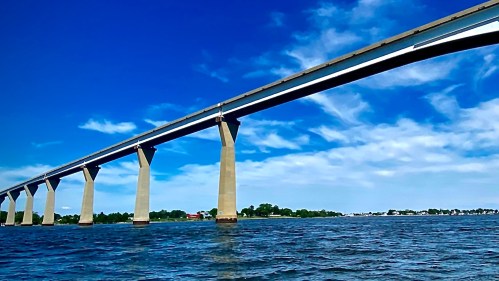 A long bridge spans over calm water, supported by tall concrete pillars, under a bright blue sky with scattered clouds.