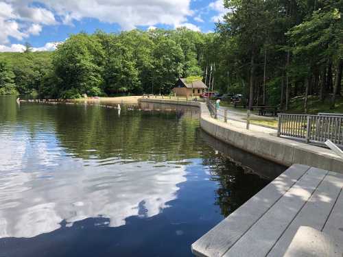 A serene lake surrounded by trees, with a path and a small building in the background under a partly cloudy sky.