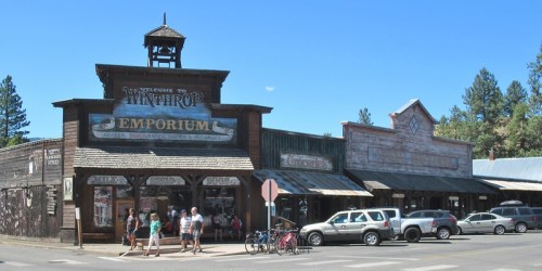 Historic buildings in Winthrop, Washington, featuring shops and a clear blue sky. People and bikes are visible in the foreground.