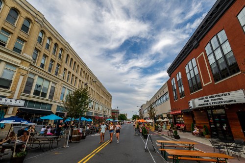 A lively street market with people walking, outdoor seating, and colorful umbrellas under a cloudy sky.