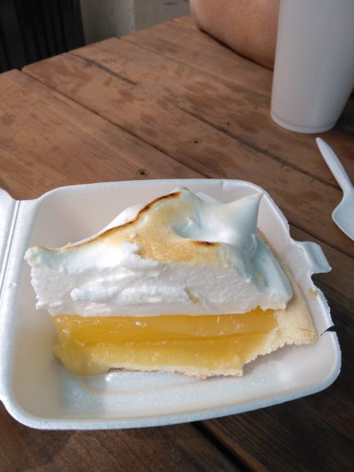 A slice of lemon meringue pie with a golden filling and fluffy white meringue on a foam takeout container.