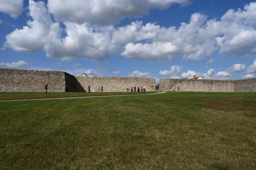 A grassy area with a stone wall in the background and people walking along a path under a cloudy sky.