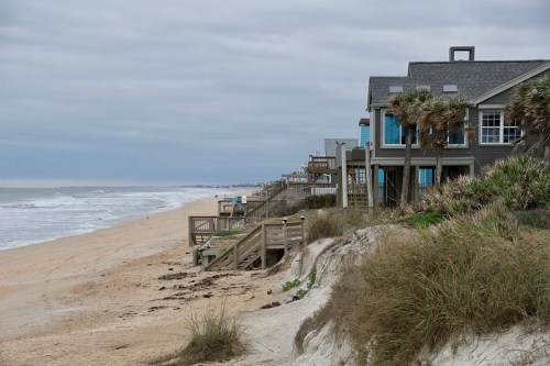 Coastal scene with beach, ocean waves, and houses along the shore under a cloudy sky.