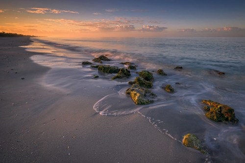 A serene beach at sunset, featuring smooth waves gently lapping over moss-covered rocks along the shore.