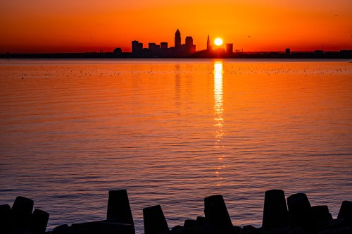 Sunset over a city skyline reflected in calm water, with silhouettes of buildings and a warm orange sky.