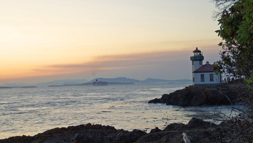 A lighthouse stands on rocky shore at sunset, with a ship sailing in the distance and mountains on the horizon.