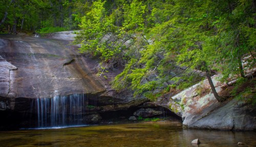 A serene waterfall cascades over rocks into a calm pool, surrounded by lush green trees and a rocky landscape.