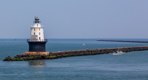 A lighthouse stands on a rocky pier, surrounded by calm blue waters and a distant boat in the background.
