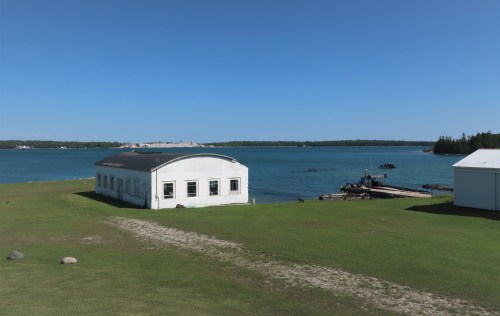 A white building near a calm blue lake, with green grass and a dock in the background under a clear blue sky.