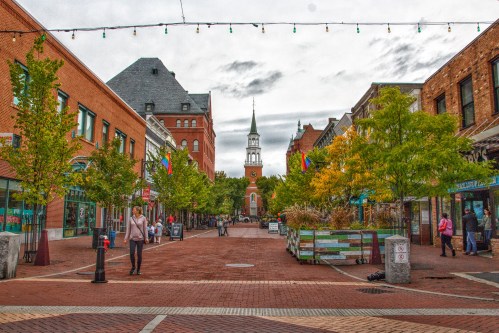 A vibrant street scene with trees, shops, and a clock tower in the background under a cloudy sky.