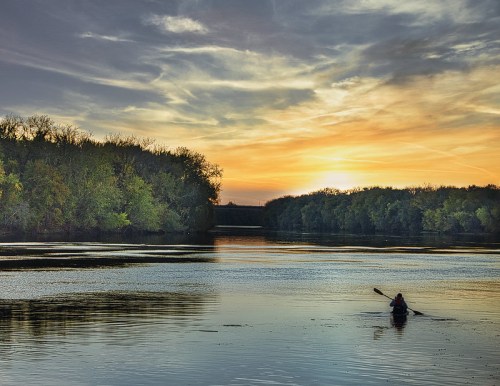 A person kayaking on a calm river at sunset, surrounded by trees and a colorful sky.