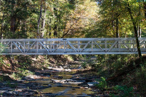 A white pedestrian bridge spans a rocky creek, surrounded by trees with autumn foliage.