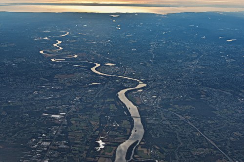 Aerial view of a winding river through a landscape of fields and urban areas, with a sunset reflecting on the water.