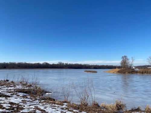 A serene river scene with a clear blue sky, snow on the ground, and a small island in the water.