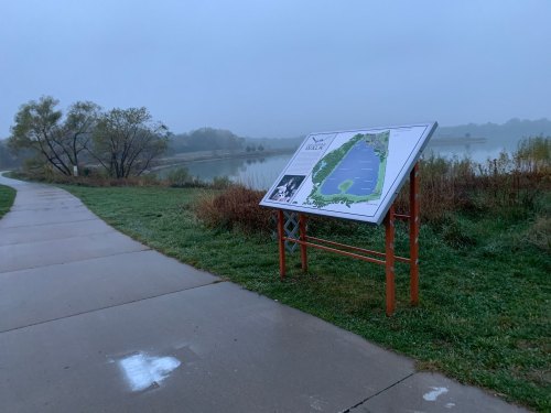 A foggy lakeside path with a map sign showing the area, surrounded by grass and trees.
