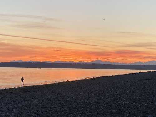 A person stands on a beach at sunset, with mountains silhouetted against a colorful sky reflecting on the water.