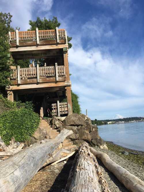 A wooden observation tower near a beach, surrounded by trees and driftwood, with a view of the water and distant mountains.