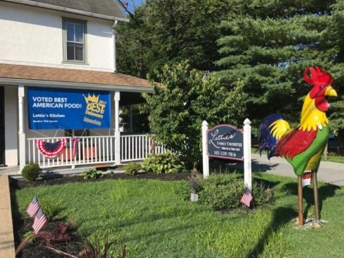 A colorful rooster statue stands in front of a restaurant with a "Voted Best American Food" banner and patriotic decorations.