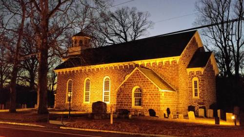 A historic stone church illuminated at dusk, surrounded by trees and a graveyard.