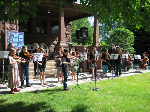 A group of young musicians playing string instruments outdoors in front of a house, with music stands and a speaker nearby.