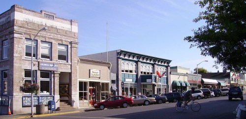 A charming street scene featuring historic buildings, shops, and parked cars under a clear blue sky.