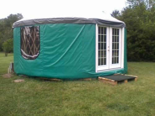 A green yurt with a gray top and double doors, set on a grassy area with a small wooden platform.