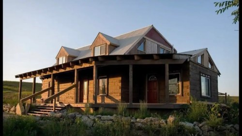 A rustic house with a wooden exterior and a porch, set against a clear sky and grassy landscape.