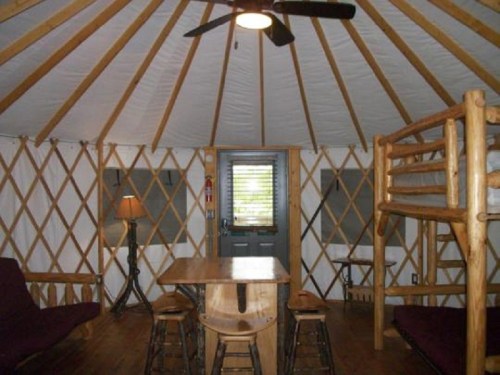 Interior of a yurt featuring wooden furniture, a table, bunk beds, and a ceiling fan. Natural light from the door.