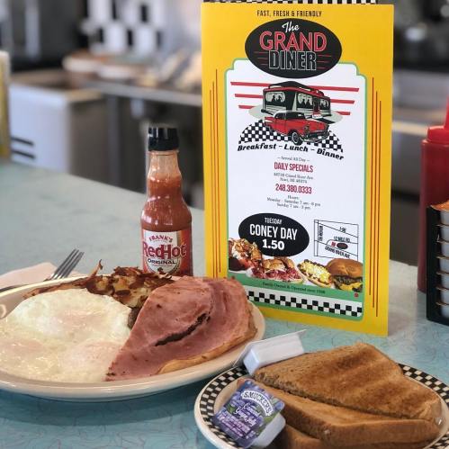 A plate of breakfast with eggs, bacon, ham, toast, and hot sauce, next to a menu for The Grand Diner.