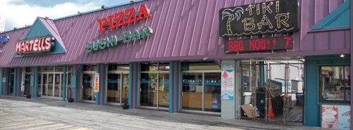 A colorful storefront featuring a pizza place, sushi bar, and tiki bar, with a purple roof and outdoor seating.