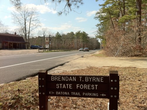Sign for Brendan T. Byrne State Forest with Batona Trail parking, alongside a road lined with trees and a clear sky.