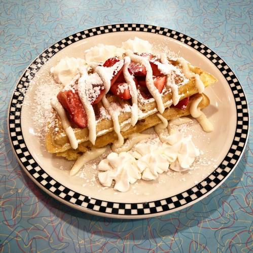 A plate of waffles topped with strawberries, whipped cream, and powdered sugar, served on a checkered rim.