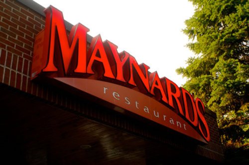 Sign for Maynards Restaurant, featuring bold red letters against a brick background, with greenery above.