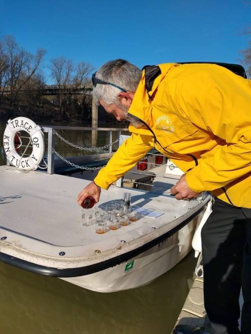A man in a yellow jacket collects samples in glass tubes on a boat by a river on a sunny day.