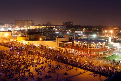 A bustling beach at night, filled with people, illuminated by lights from nearby buildings and restaurants.