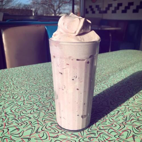 A tall glass of pink milkshake topped with whipped cream, set on a retro-patterned table in a diner.