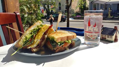 A plate with a sandwich stacked with vegetables and bacon, alongside a glass of water, on a sunny outdoor table.