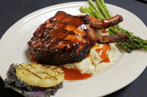 A grilled pork chop with barbecue sauce, served with mashed potatoes, asparagus, and a grilled pineapple slice.