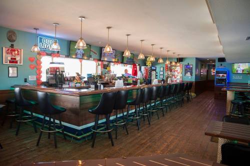 A brightly lit bar with high stools, colorful decor, and a long wooden counter, featuring various drink signs.
